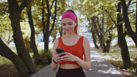 Athletic-fitness-sport-runner-girl-walking,-using-smartphone-listening-music-with-earphones-in-park