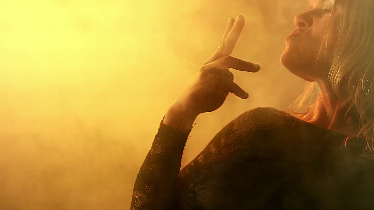 alegre chica rubia elegante con cabello corto bailando y haciendo acción de fumar con los dedos en el humo y la luz de neón