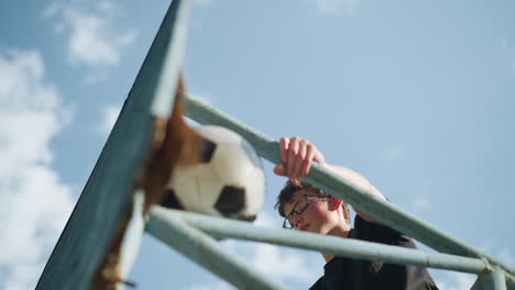 an upward view of a young boy sitting on outdoor blue iron equipment, holding the iron with his left hand while adjusting his glasses with his right hand, a ball is lodged within the iron