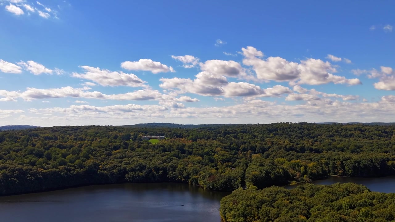 Aerial tilted hyper lapse over the Muscoot Reservoir surrounded by trees and pristine natural habitat. The camera dolly in and roll left over the horizon on a sunny day with blue skies and clouds.