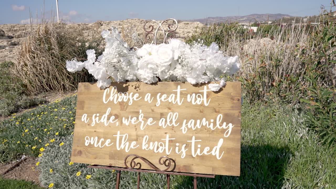 Un cartello dice: scegli un posto, non una fazione; siamo tutti una famiglia una volta celebrato il matrimonio. L’ambientazione è un giardino con fiori e vegetazione durante un matrimonio.