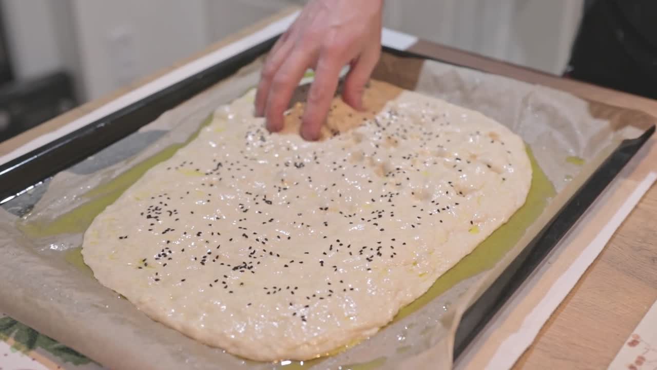 Un amatore che prepara la focaccia. Mani che premono e versano olio d'oliva sulla forma. Tutorial sulla preparazione della focaccia. Messa in risalto della formazione di bolle in superficie prima della cottura. Contenuti visivi autentici di cucina casalinga