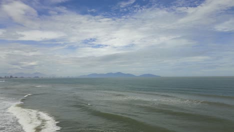 waves in the ocean breaking towards the coast with big fluffy clouds in the sky and mountains in the horizon