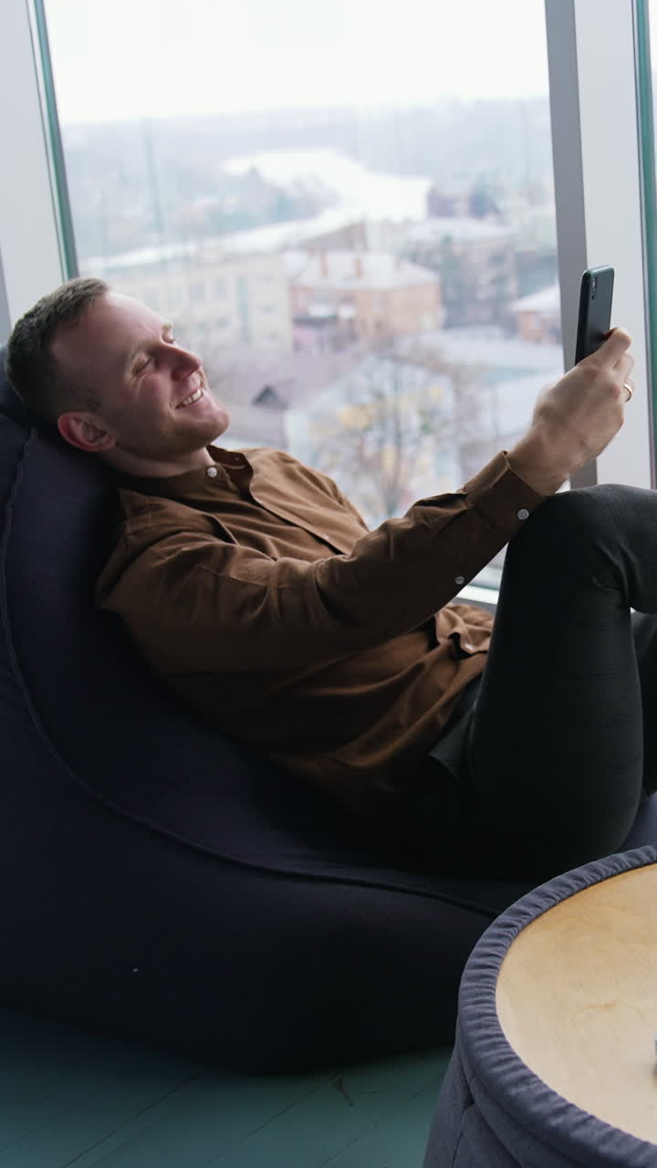 Young businessman relaxing near the window. Handsome man sitting in a cosy chair and having a video conversation with the city window view. Profile view. Vertical video
