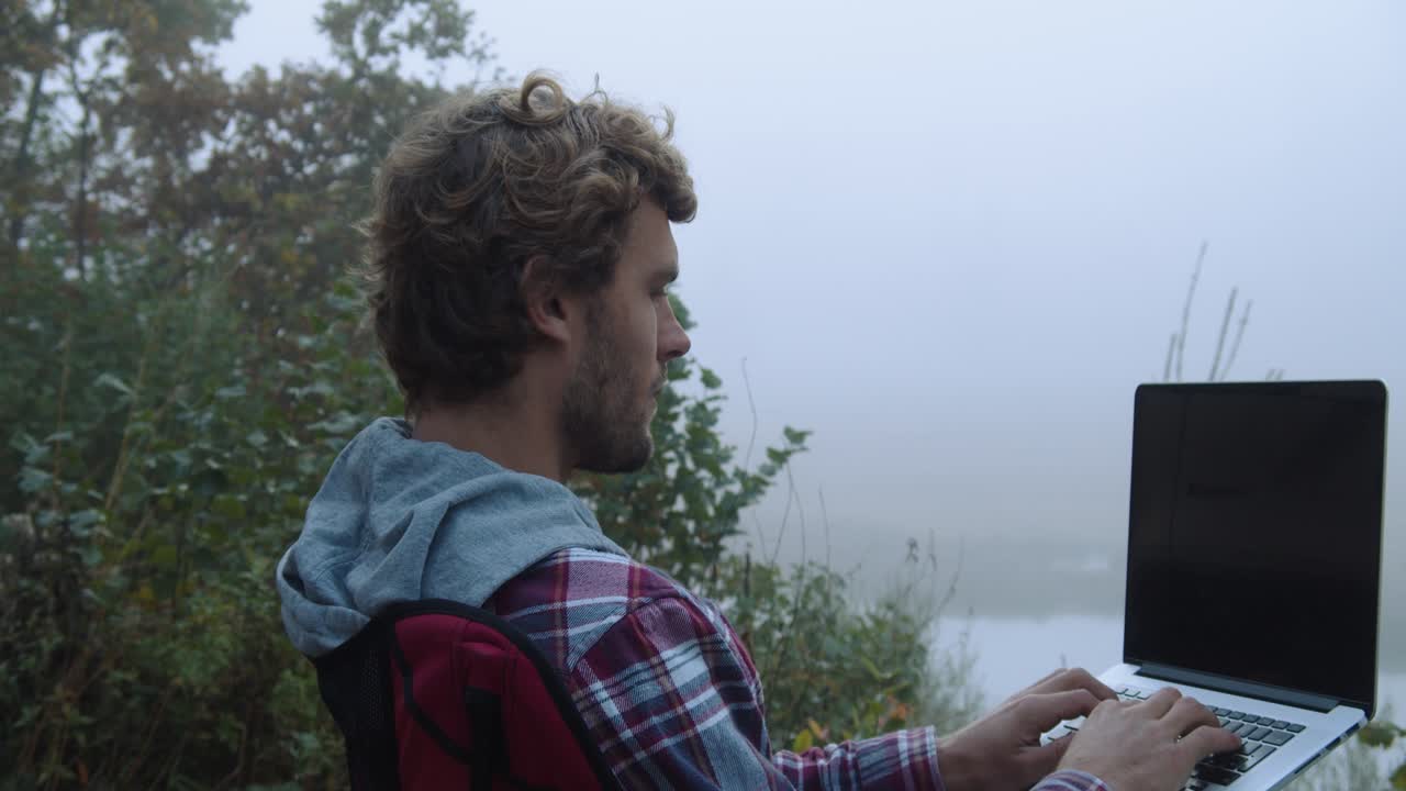 Close up shot, a man using a laptop, foggy trees and lake in the background