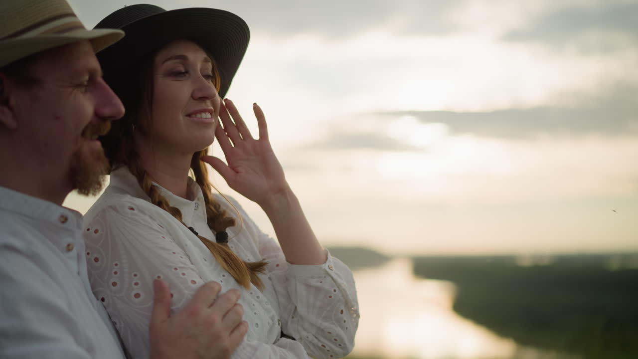 A loving couple dressed in white shirts and hats embraces tenderly at sunset. The man holds the woman from behind as she points into the distance, sharing a moment of connection and exploration