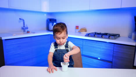 beautiful baby boy in jeans romper sits at the kitchen table. cute toddler eats yogurt with a spoon.