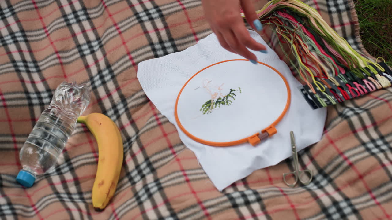 Close up of person picking embroidery thread from colorful skeins on patterned blanket beside water bottle, banana, and scissors, showing calm outdoor crafting scene filled with creativity