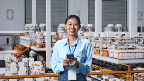 una mujer de negocios asiática usando un teléfono móvil y sonriendo a la cámara en el almacén