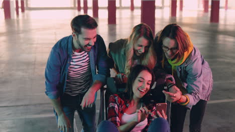 grupo caucásico de amigos viendo algo en el smartphone y riéndose mientras dos de las chicas están sentadas en un carrito en un parque vacío