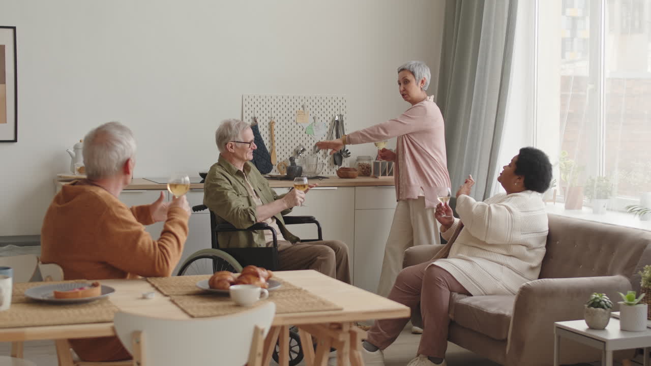 Steadicam of African woman sitting on couch, Asian lady standing near, grey-haired person at table and Caucasian man in wheelchair smiling, having conversation, drinking white wine