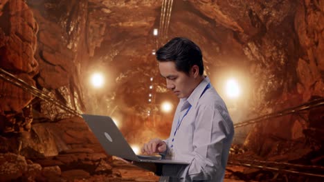 side view of an asian male professional worker standing with his laptop in underground mine tunnel, typing on his laptop's keybaord with meditation