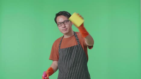 sonriente ama de casa masculina asiática con un delantal lavando vidrio por un spray y esponja de cocina en el estudio de pantalla verde