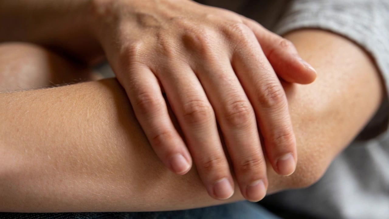 La mano derecha de una persona se acomoda en el regazo, haciendo pequeños ajustes en el antebrazo izquierdo en casa, vistiendo una camiseta gris.