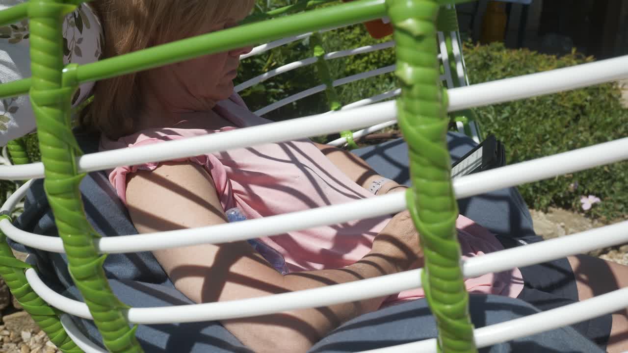 편안한 정원 의자 그네를 탄 여성은 화창한 날 정원에서 편안한 전자책 소설을 읽습니다.