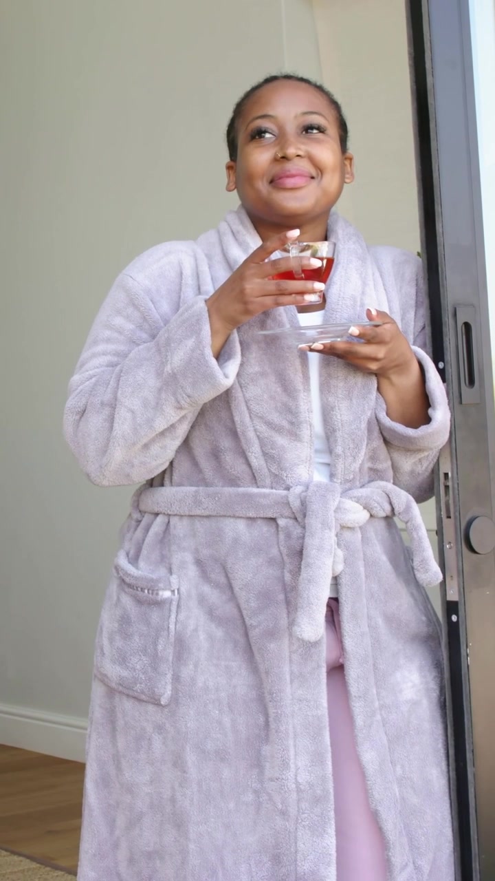 Vertical video: Sipping woman in gray robe with glass teacup and saucer at sliding doorway, smiling