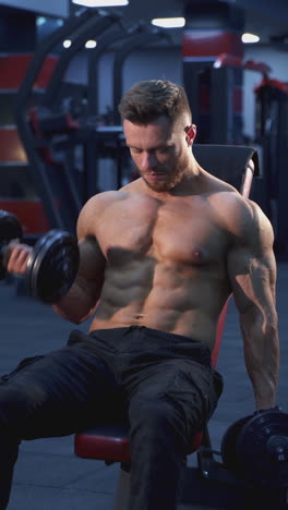 professional bodybuilder exercising in gym. shirtless muscular athlete training his muscles with dumbbells while sitting on chair in sports center. vertical video