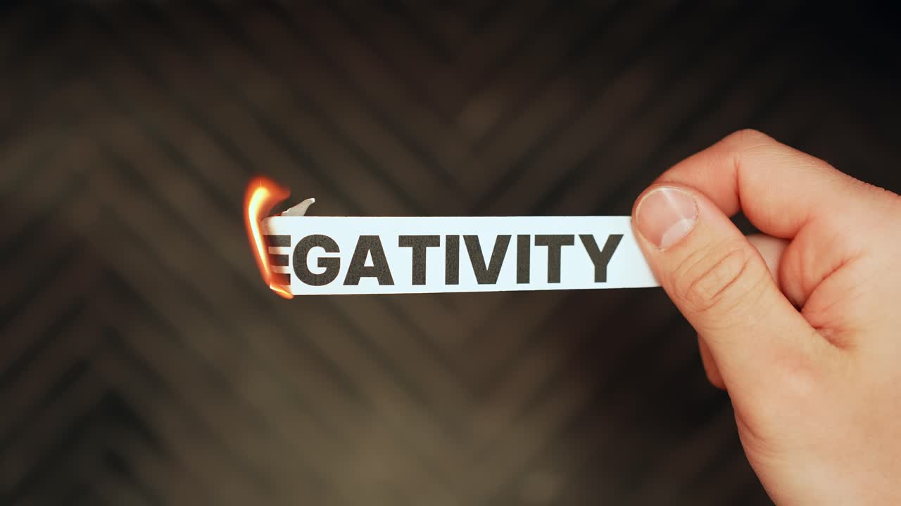Close-up of a hand holding a paper strip labeled “NEGATIVITY” as the edge burns with flame on a dark background. Letting go of toxic thoughts concept with dramatic copy space