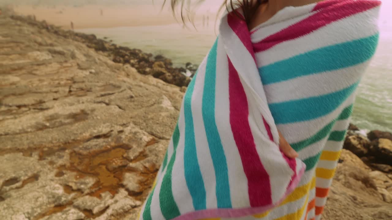 Ung kvinna insvept i en randig handduk står på en klippig strand