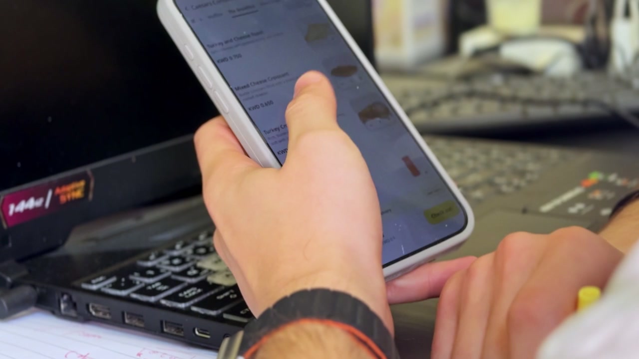 Close-up of a person holding a smartphone beside an open laptop in an indoor office workspace, highlighting mobile technology, computer use, and everyday digital work.