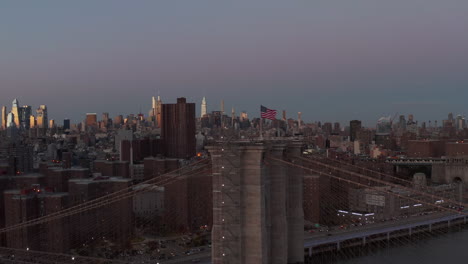 Vuela-Alrededor-De-La-Torre-De-Apoyo-Del-Puente-De-Brooklyn-Con-La-Bandera-Estadounidense-Levantada-Al-Atardecer.-Edificios-Altos-Iluminados-En-La-Distancia.-Manhattan,-Ciudad-De-Nueva-York,-Estados-Unidos