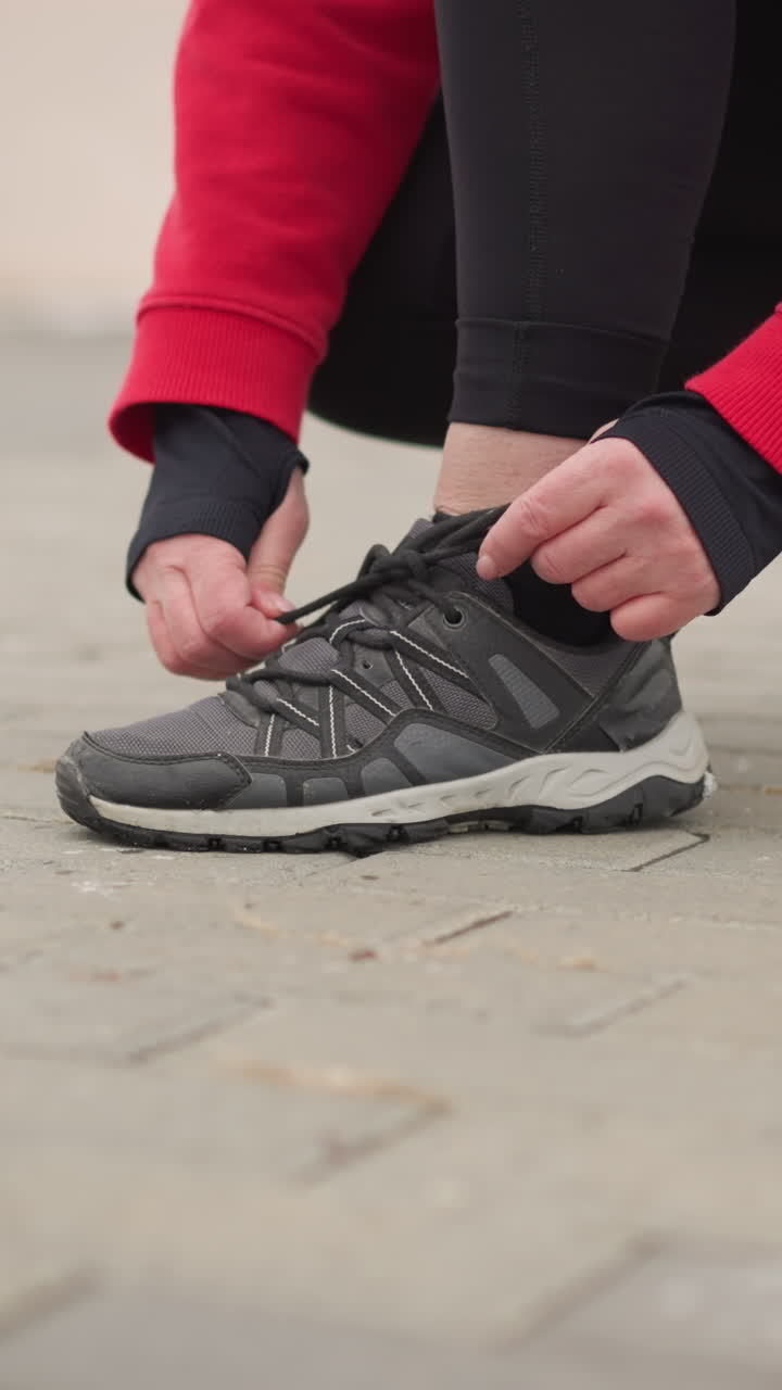 vista laterale di un'atleta che si lega il laccio della scarpa destra mentre si accovaccia su un sentiero interconnesso con uno sfondo nevoso sfocato, raffigura la preparazione concentrata per un'attività all'aperto in inverno