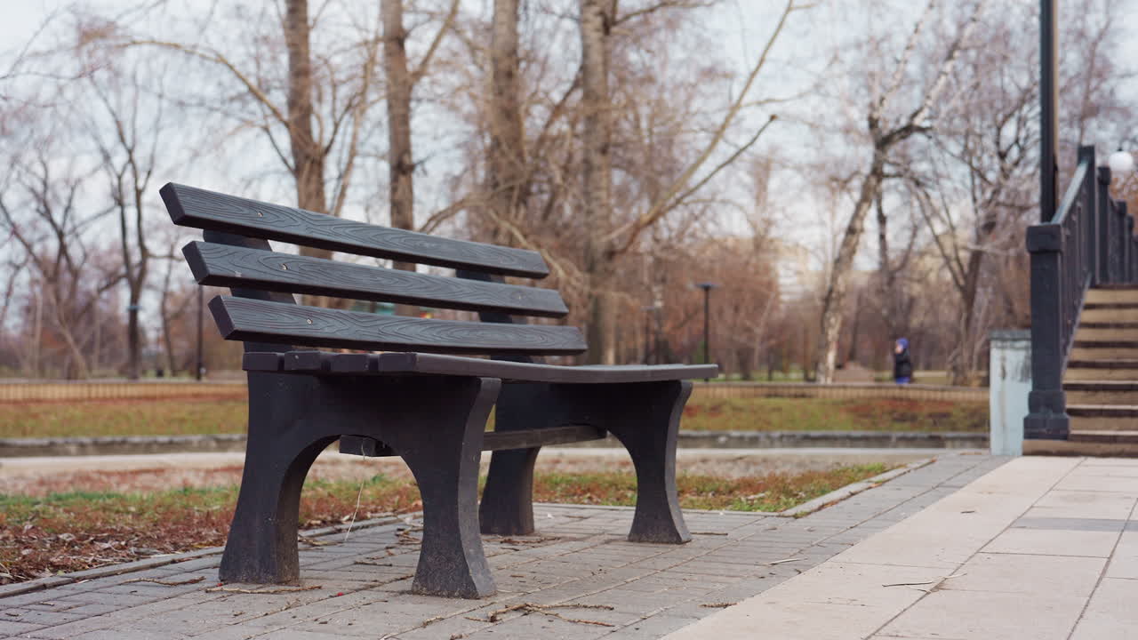 Empty wooden bench positioned near pedestrian bridge staircase in quiet winter park surrounded by bare trees, paving tiles, and soft natural tones creating calm outdoor environment
