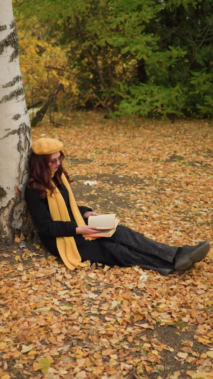 노란 베레를 입은 젊은 여자가 밖에서 앉아 나무에 기대어 은 가을 잎으로 둘러싸인 책을 고, 자연 속에서 평화로운 고독을 즐기고, 생각에 빠져, 독서에 몰두하고 있습니다.