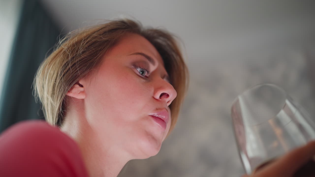 Upward view of lady in pink shirt drinking red wine as soft light reflects off face and glass showing serene relaxed moment in cozy indoor setting with calm expression and gentle natural lighting