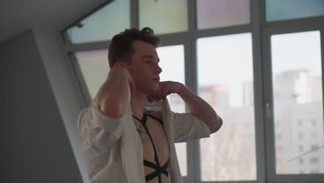 young man wearing black straps pulls on white shirt in front of large window with urban buildings outside, natural daylight casting soft glow over interior space during clothing adjustment moment