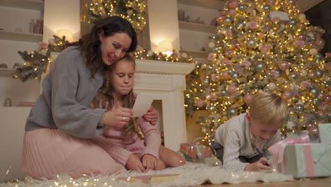 niño y niña escriben y leen una carta con su madre en el suelo junto a los regalos y el árbol de navidad