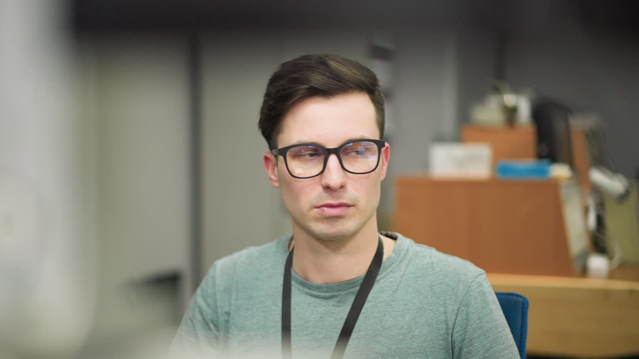Young man with glasses staring intently at computer screen in modern office setting. Focused on work, professional environment, with attention to detail and task concentration