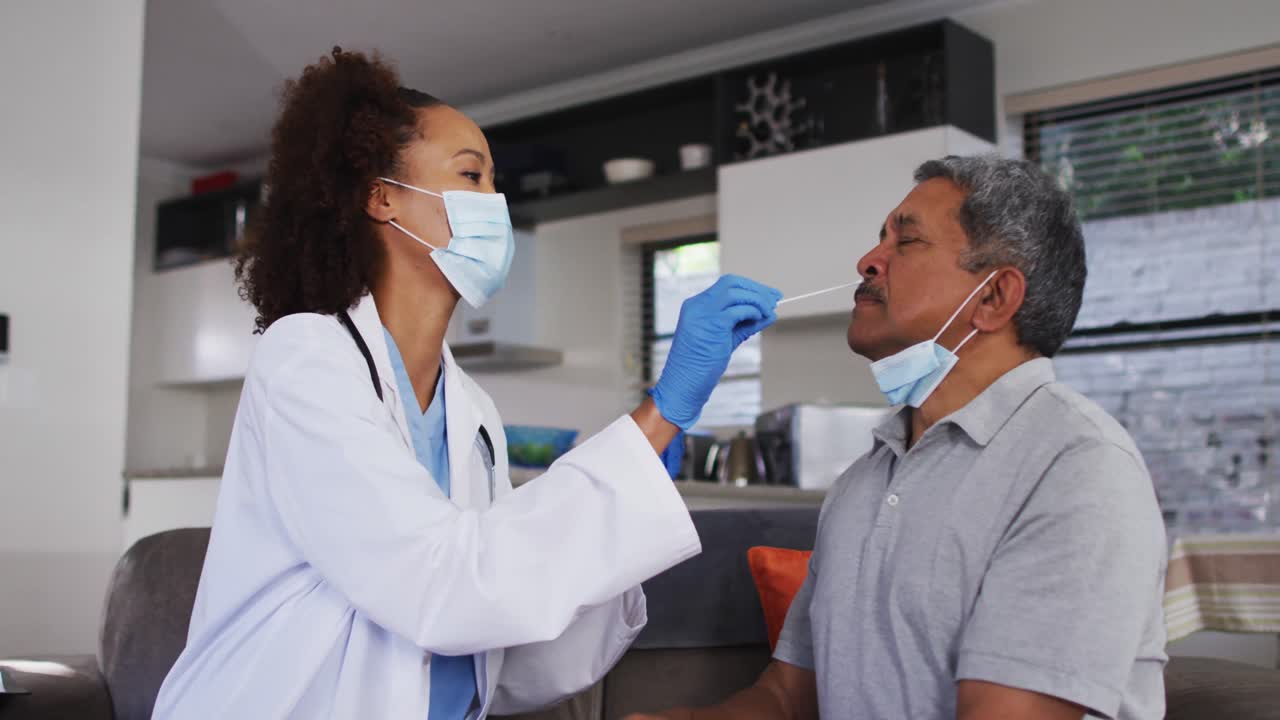Mixed race female doctor wearing mask doing swab test on senior man at home