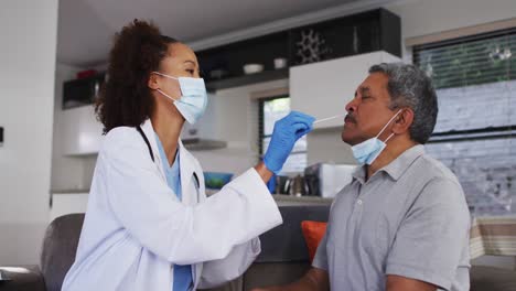 Mixed-race-female-doctor-wearing-mask-doing-swab-test-on-senior-man-at-home