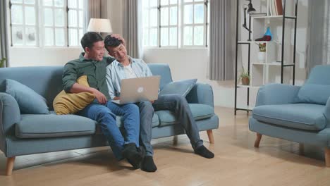 asian gentle gay couple using laptop computer, while sitting on a couch in cozy stylish apartment. adult boyfriends online shopping on internet, watching funny videos on streaming service
