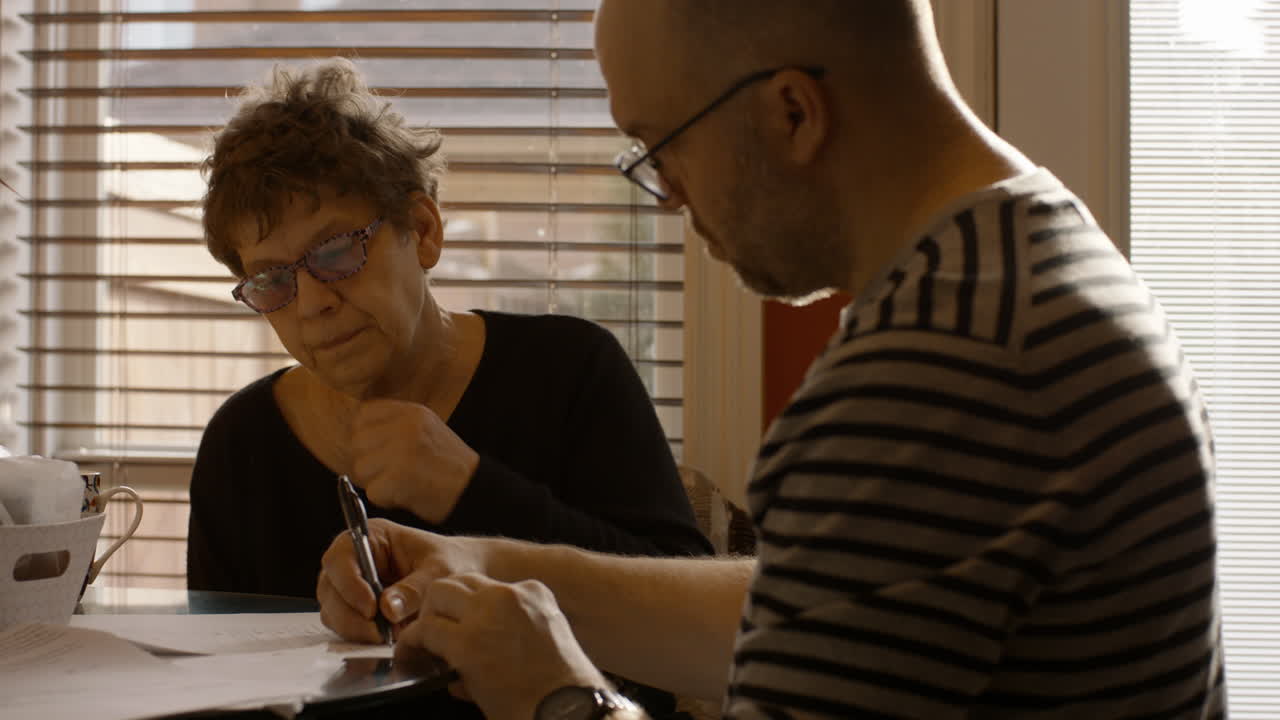 An elderly woman sits at a kitchen table with a younger man. They are reviewing, and signing documents together. The man signs first, followed by the woman. Both wear reading glasses. Warmly sun lit.