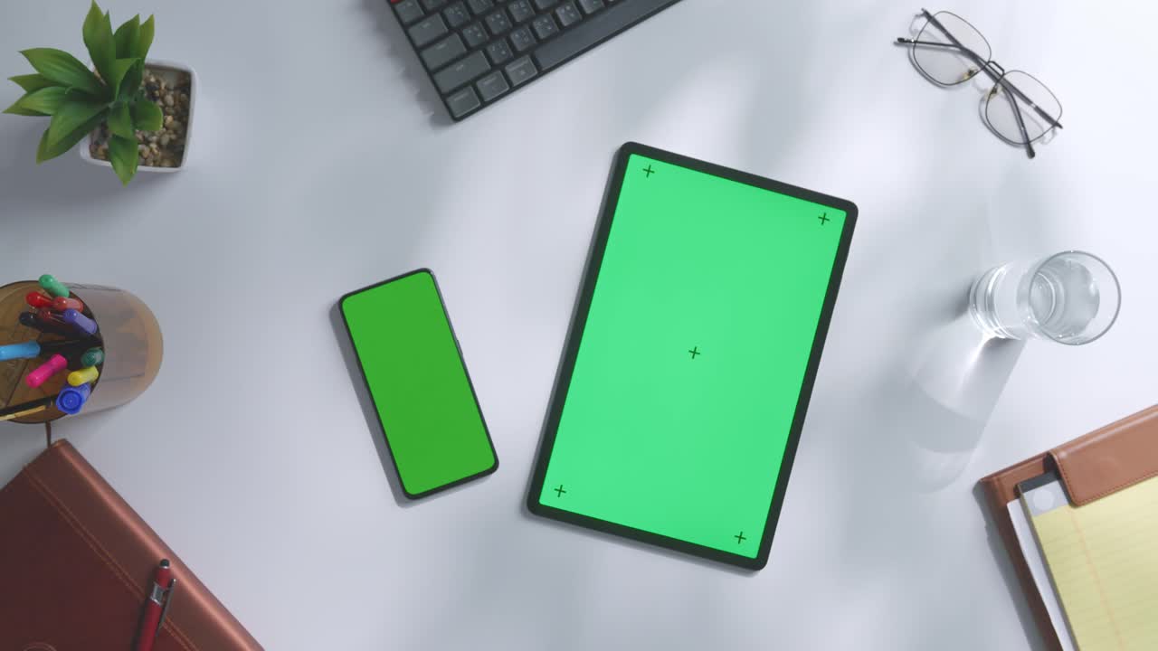 верхний вид ноутбука с макетным зеленым экраном chromakey на деревянном офисном столе рядом с ноутбуком с ручками, очками и стаканом воды.