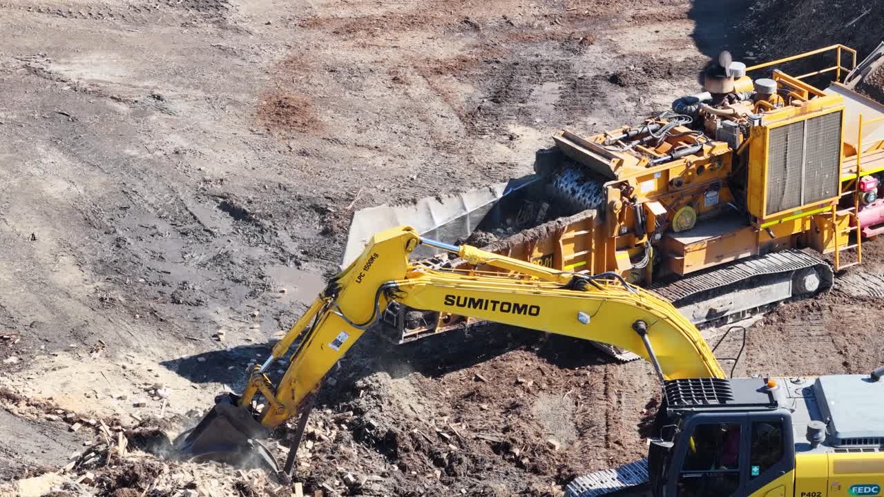 An excavator transfers soil into a processing machine in a sunlit, industrial landscape. Captured from a drone perspective