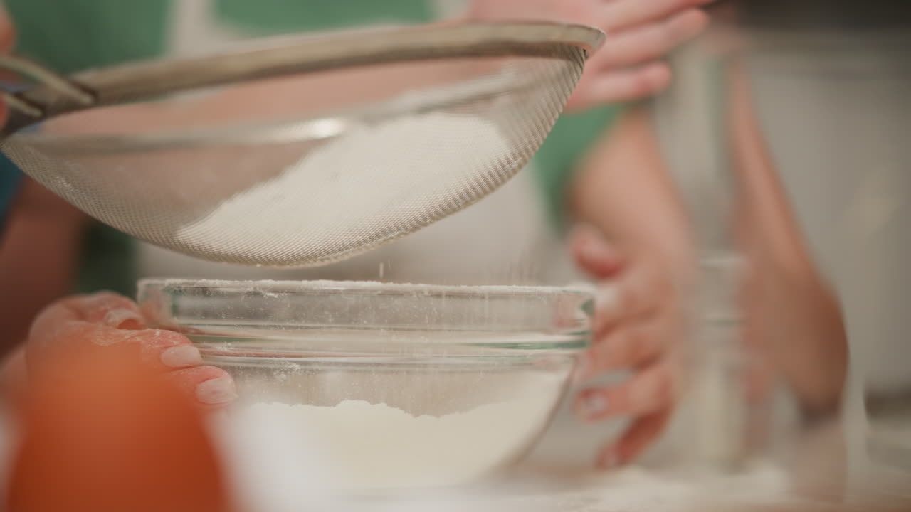 close up of person supporting glass bowl as another person carefully sieves white flour into it on kitchen surface, surrounded by baking tools and ingredients with soft blur in background