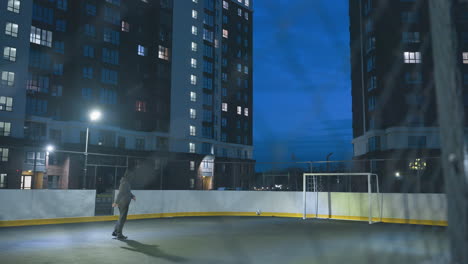 athlete seen through entrance kicks soccer ball toward goal post under bright stadium lights on urban field, framed by tall residential buildings and vivid blue night sky