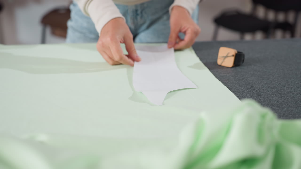 Clothing designer carefully picks up paper pattern to adjust on light green fabric spread over workspace table, preparing textile for cutting in bright creative sewing studio filled with materials