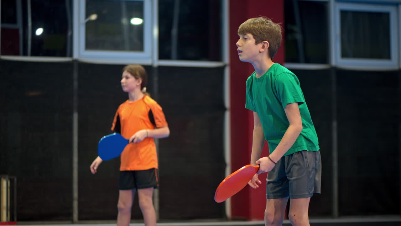 A boy and a girl playing pickleball on a blue, inside court