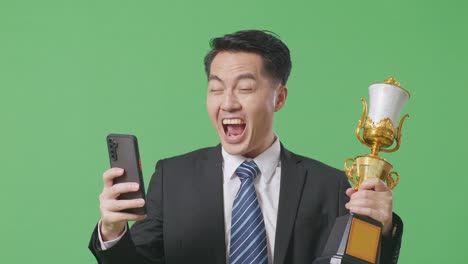 close up of asian business man in a suit and tie with a gold medal and trophy looking at smartphone then screaming goal celebrating on green screen background in the studio