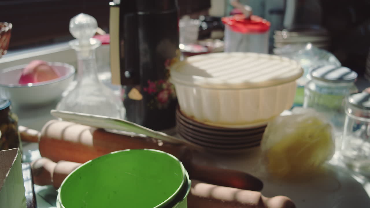 A person's hands place utensils, bowls, mixers and other kitchen utensils on a sunlit kitchen counter