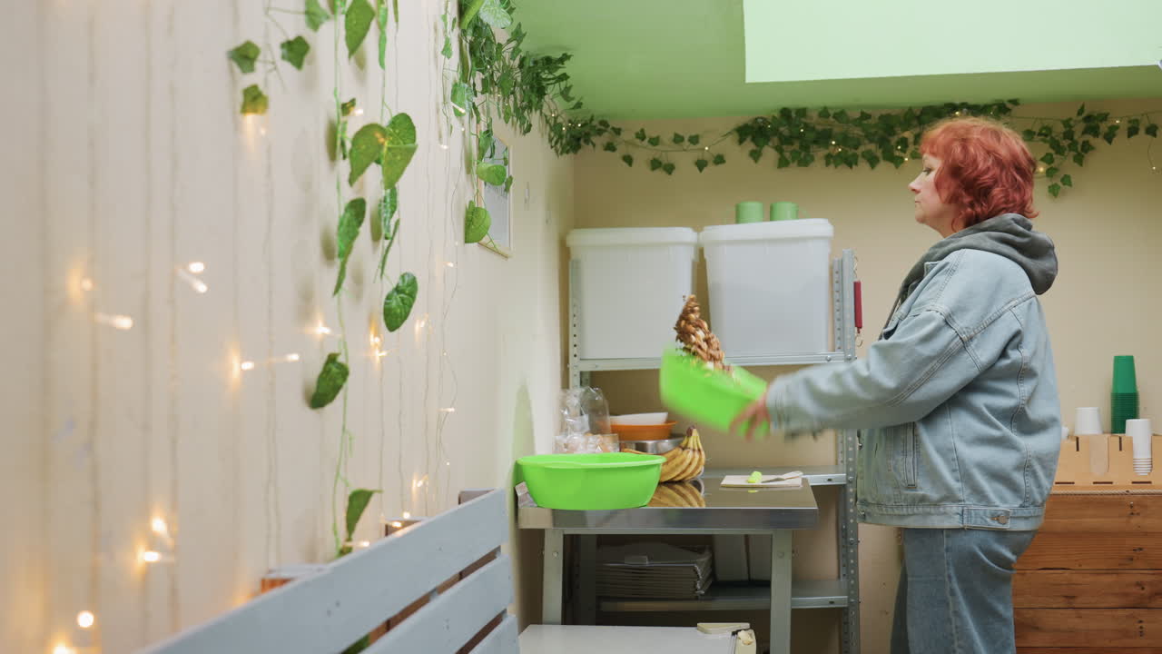 Donna bianca prepara ciotole verdi al bancone, studio decorato con piante, edera e lucine, giacca di jeans, scaffali con barattoli e packaging ecologico, mani delicate che preparano gli ordini, calma e concentrata