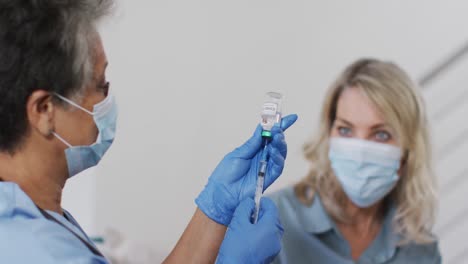 senior african american female doctor preparing vaccination for woman at home wearing face mask