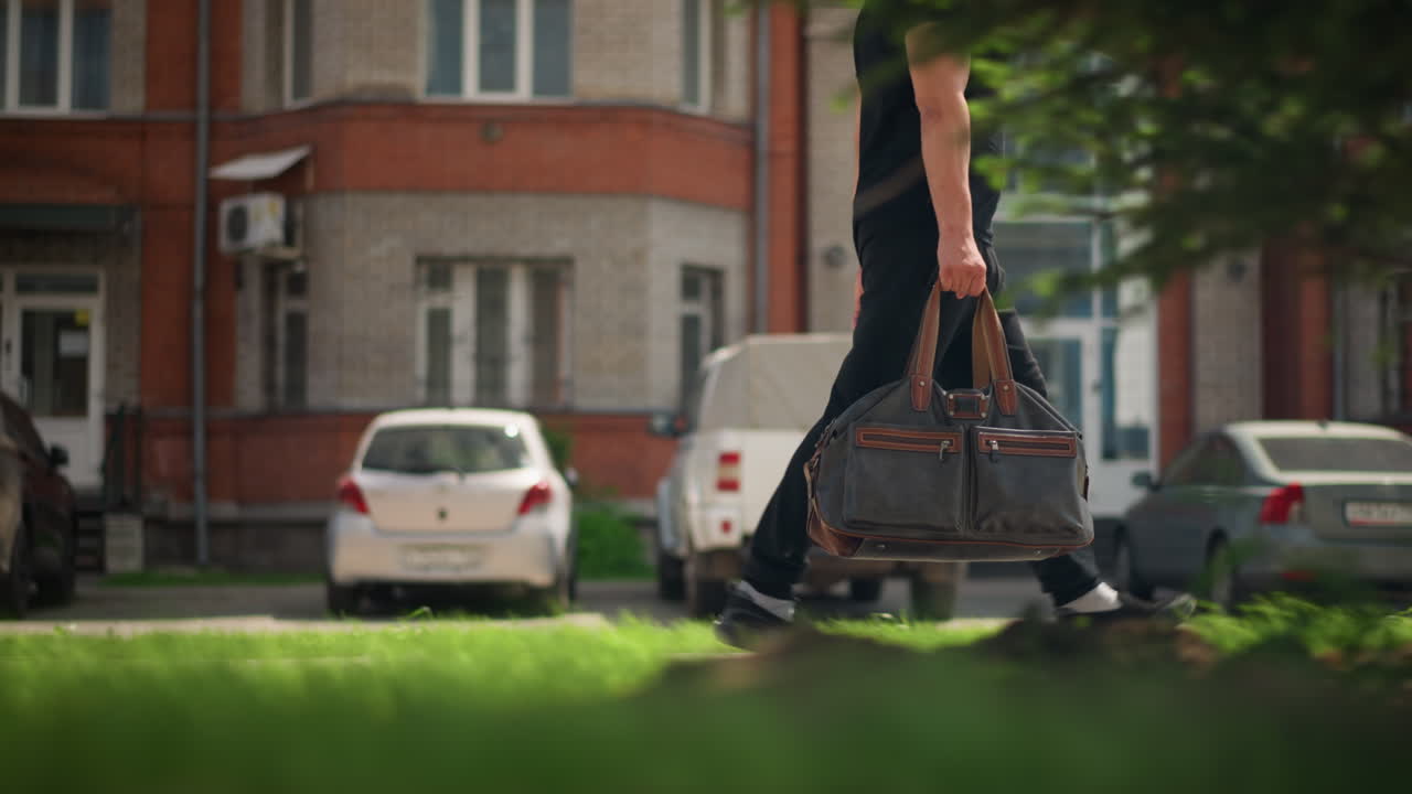 革のバッグを持った男性、屋外を歩く男性のクローズアップ、晴れた通りで革のバッグを運ぶ男性のクローズアップ、都市の駐車場脇の晴れた舗道を革のバッグを持って歩く男性。