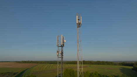 Two-radio-towers-in-the-middle-of-farmland-during-sunrise,-aerial-tilting-downward