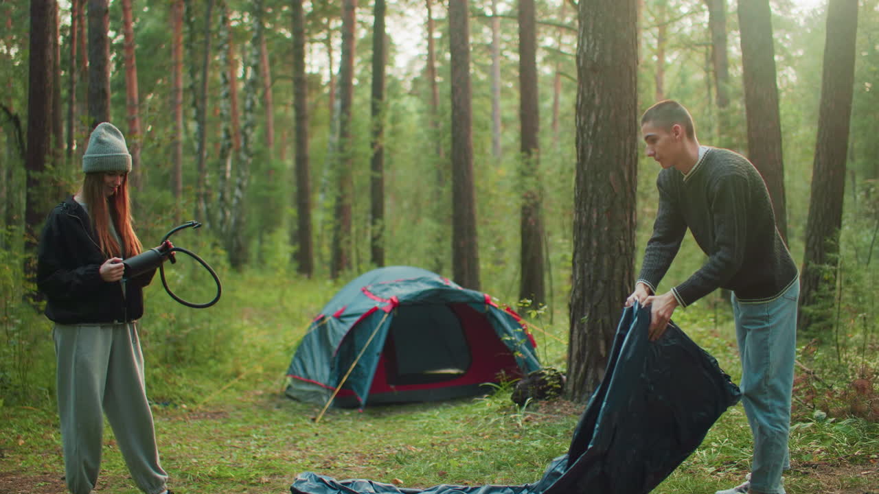 Young woman holding air pump stands in forest clearing watching man spray tent fabric beside trees and camping gear, sunlight filtering through woods as couple prepares outdoor campsite