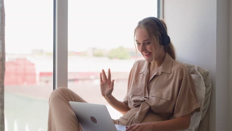 young woman waves hand at videocall via laptop on windowsill